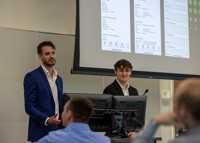 Two individuals presenting at a conference in front of a large screen displaying a PowerPoint presentation.
