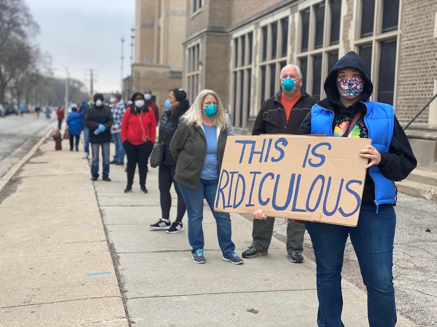 Voters standing in line in Milwaukee, wearing masks and holding a cardboard sign saying “THIS IS RIDICULOUS”