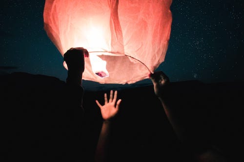 A hand reaching up towards a lit sky lantern at night, with stars in the background.