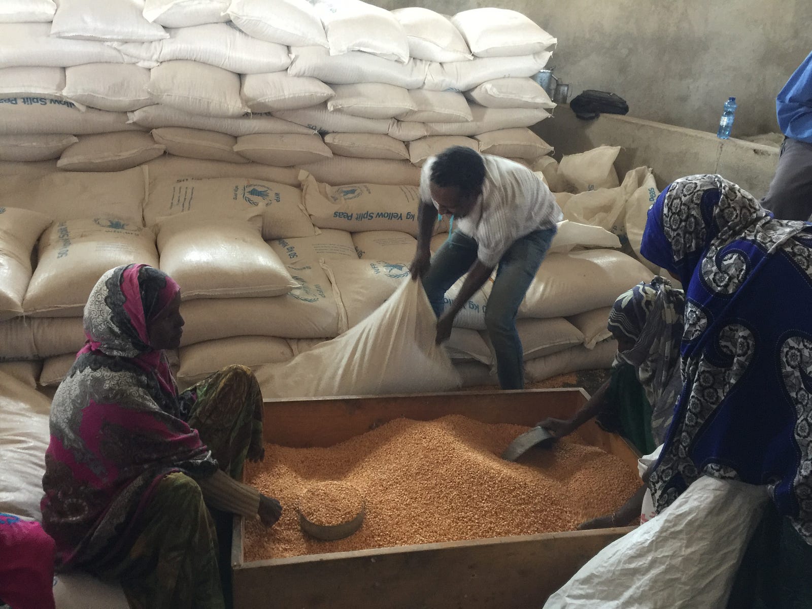 World Food Program in Ethiopia An African man and woman kneel next to a box of grain, behind them are large white bags of grain, one man is lifting a bag.