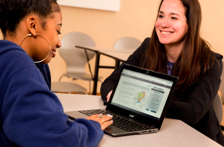 A teacher and student sitting across from each other at a table, smiling. The student is working on a CommonLit lesson.
