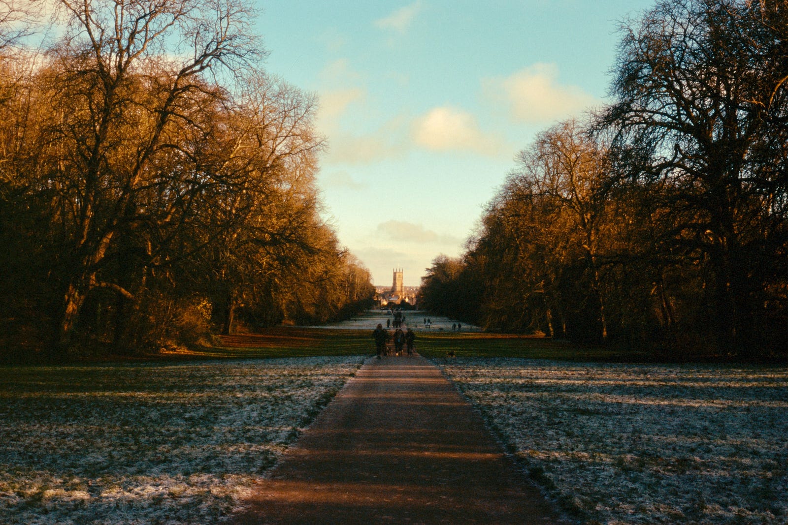 A colour photograph depicting a view in Cirencester Park looking towards the Abbey. Snow is dusted on the ground, and the sky is a wan colour of blue, with some light, puffy clouds in the sky.