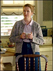 A woman wearing a denim skirt and grey cardigan stands by a kitchen counter holding a cup of tea.