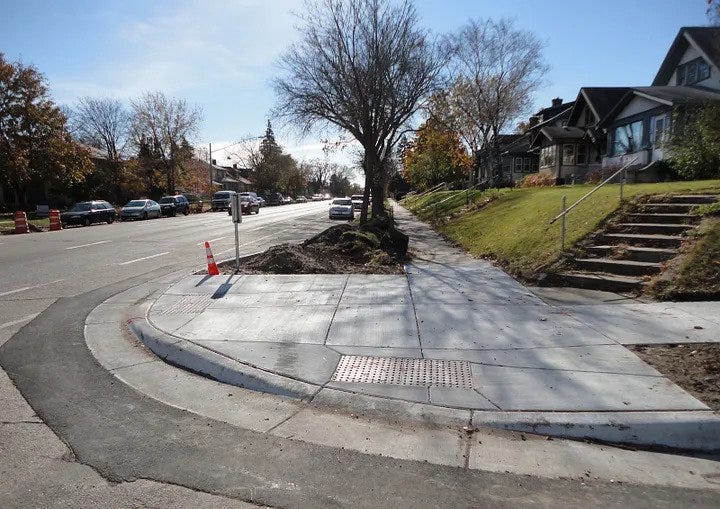 Corner of suburban street with curb cut ramp up to sidewalk. The first house has stairs to the entrance.