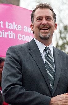 An upclose shot of Washington State Senator Kevin Ranker in front of a pink sign. Ranker is wearing a gray suit, white shirt,