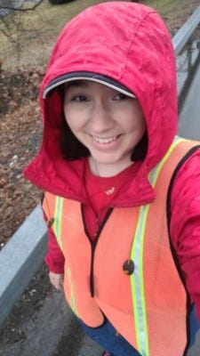 The author walking down the street in the rain. Wearing a red rain coat with the hood up and an orange reflective vest.