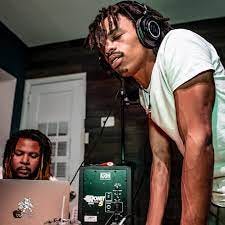 YG Marley stands, leaning on a table wearing headphones, with his eyes closed as he appears to listen to music. Another man sits in front of an open laptop. They both appear to be working on creating music.