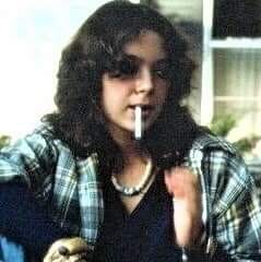 Diane wears a flannel, black tank top, and a necklace. Her hair is shoulder-length, wavy, and she holds a cigarette in her mouth, while looking down.