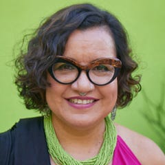 Bright green background behind a close-up of Martha, who is wearing thick-rimmed glasses and a green, pink, and black top. Her hair is in a curly bob.