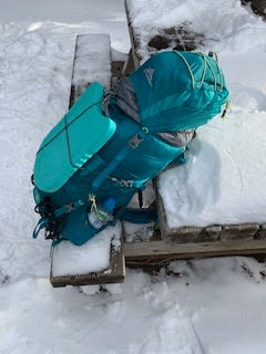 A blue backpack with straps tied down and a flat kickboard (snow seat) strapped to the front with small bungee cords. This sits on a snowy picnic table that is the background for this picture.