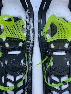 A close up of two snowshoes with bright green/yellow binding and black bases. They both have light, faint writing on the center of the shoe: “left”. There is snow in the background of the picture that they are resting on.