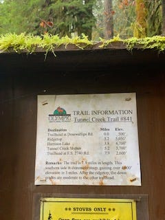 A moss-covered brown shelter for a small white square of paper with trail information. Sign says: Trail Information Tunnel Creek Trail #841 Trailhead at Dosewallips Road: Miles 0.0 Elev. 500' Ridgetop: Miles 3.2 Elev. 5050' Harrison Lake Miles 3.8 Elev 4,700' Remarks: The trail is 7.9 miles in length. This southern side is extremely steep, gaining over 4,000' elev in 3 miles.