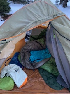 A close up of the inside of an open tent taken from the outside. From left to right: green dry bag, gray dry bag (sea to summit), a green, open sleeping quilt with a blue liner, a (deflated) sleeping pad, and the rest of the tent flap. In the background is barely visible snow with the bottoms of a few trees