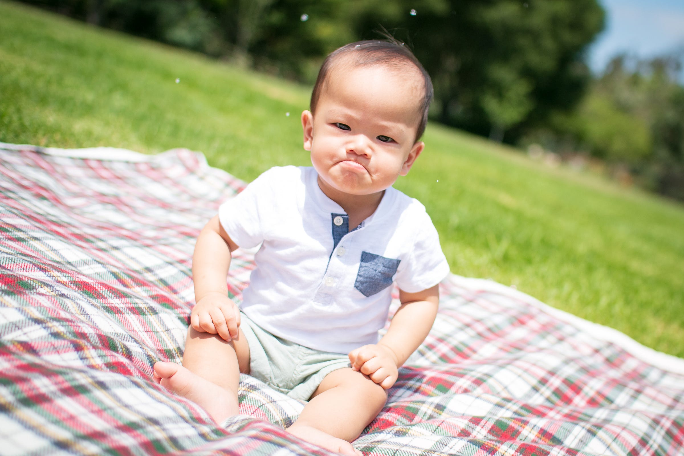 a baby with downturned lips sitting on a plaid blanket on grass