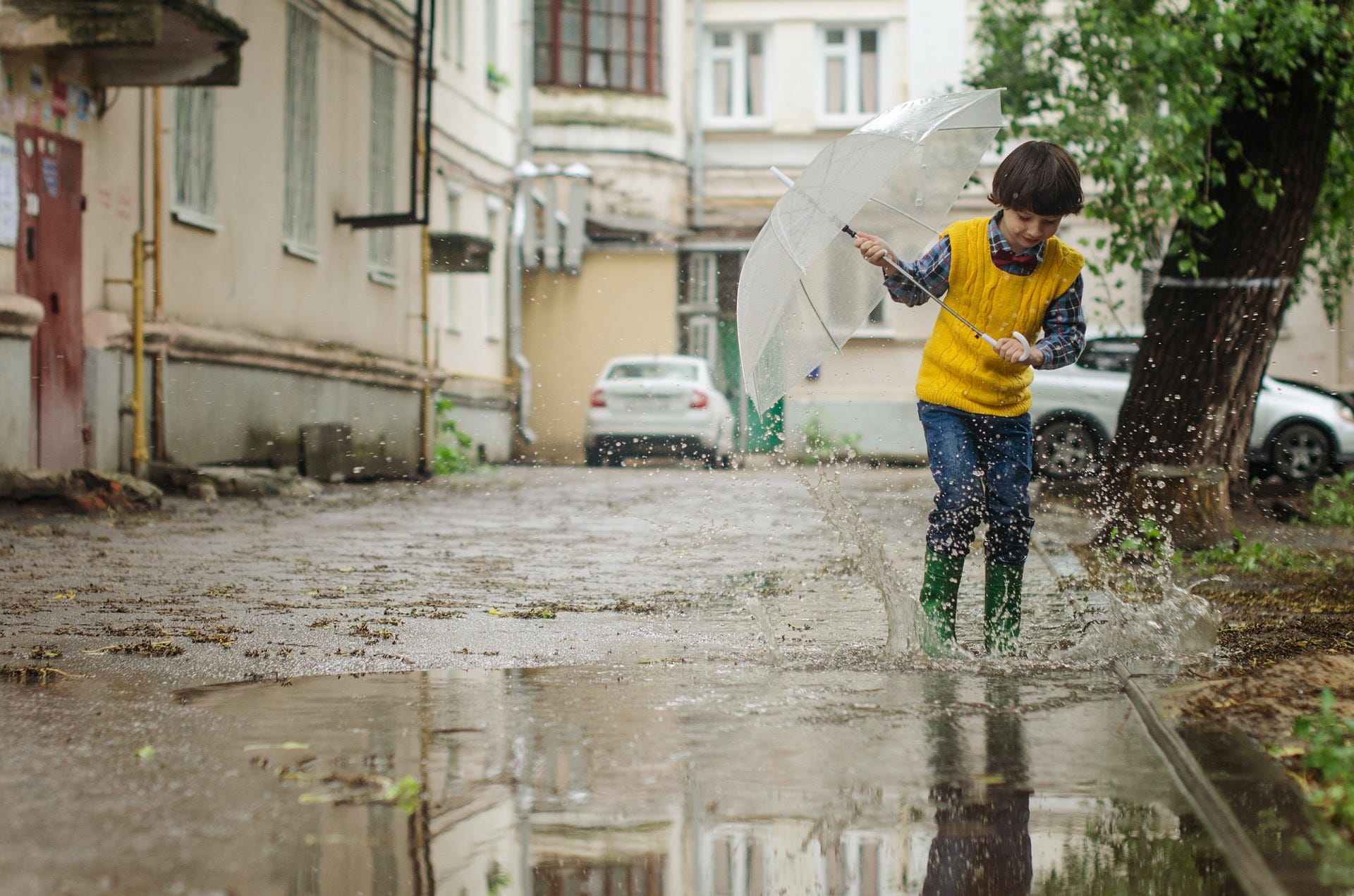 A happy boy with umbrella jumping in a puddle of water in a street