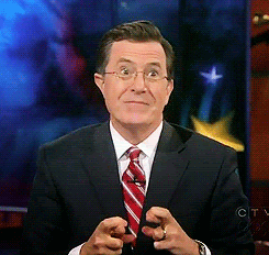 Stephen Colbert awkwardly smiling and crossing his index and middle fingers on both hands.