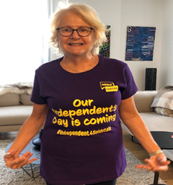 Vicky Cogley stands smiling at the camera pointing to her purple t-shirt with gold text that reads ‘Out independants day is coming’.