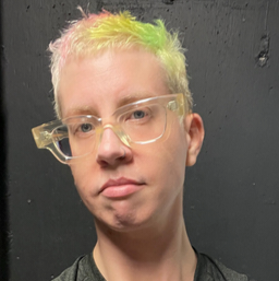 Doctor Phoenix Andrews has short cropped hair that is stripped with neon green, yellow and orange, and is wearing clear plastic rimmed spectacles and a dark grey t-shirt, whilst standing in front of a black painted wall.