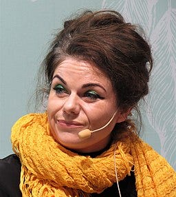 Caitlin in front of pale wall, looking to right, wearing sparkly green eyeshadow, winged eyeliner and a large yellow scarf