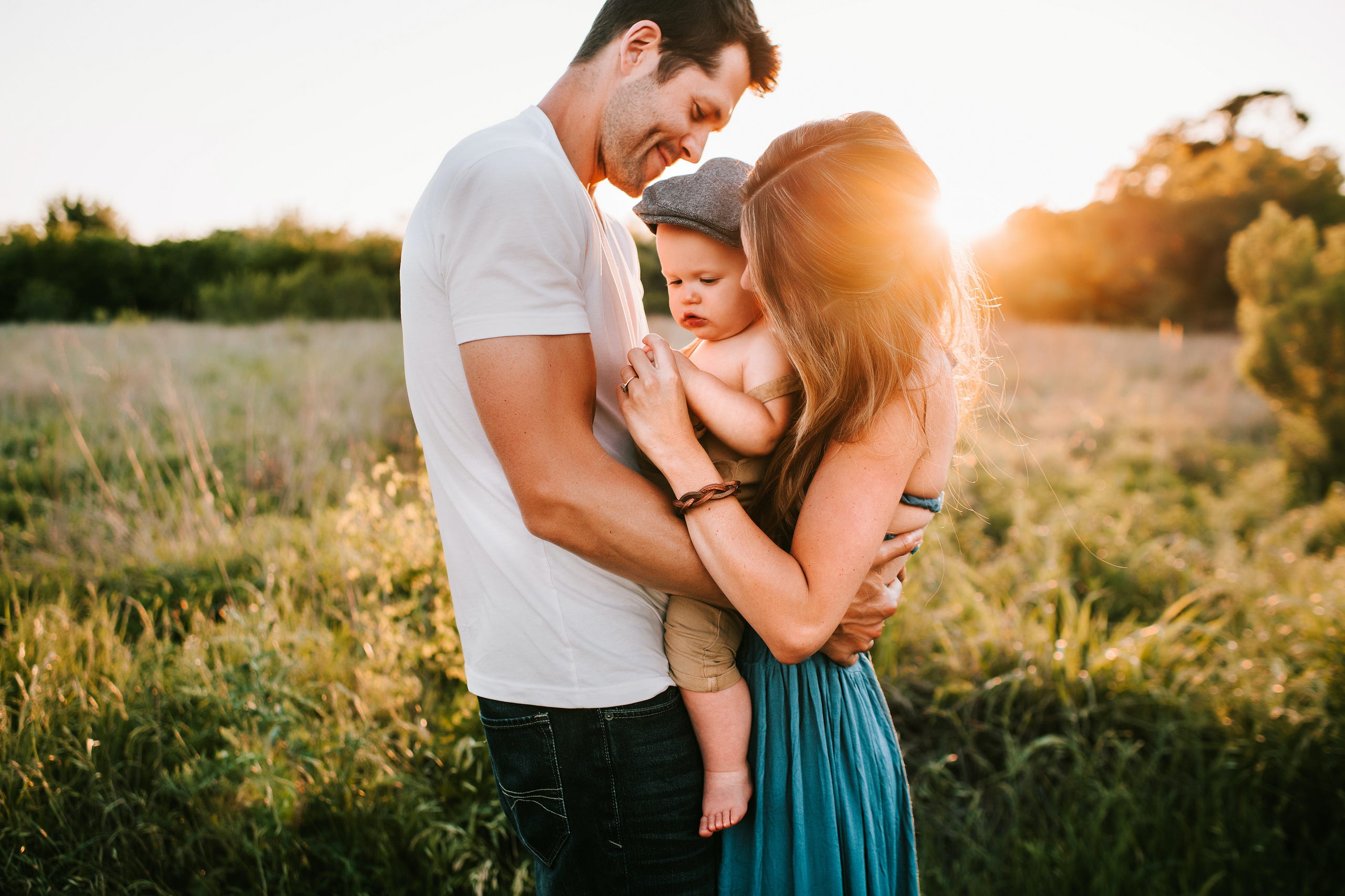 In a field in the background with a sun setting below the background tree-lining the fields are two new parents embracing one another lovingly with a young infant baby wearing a cap and no clothes in between them and being played with by his parents