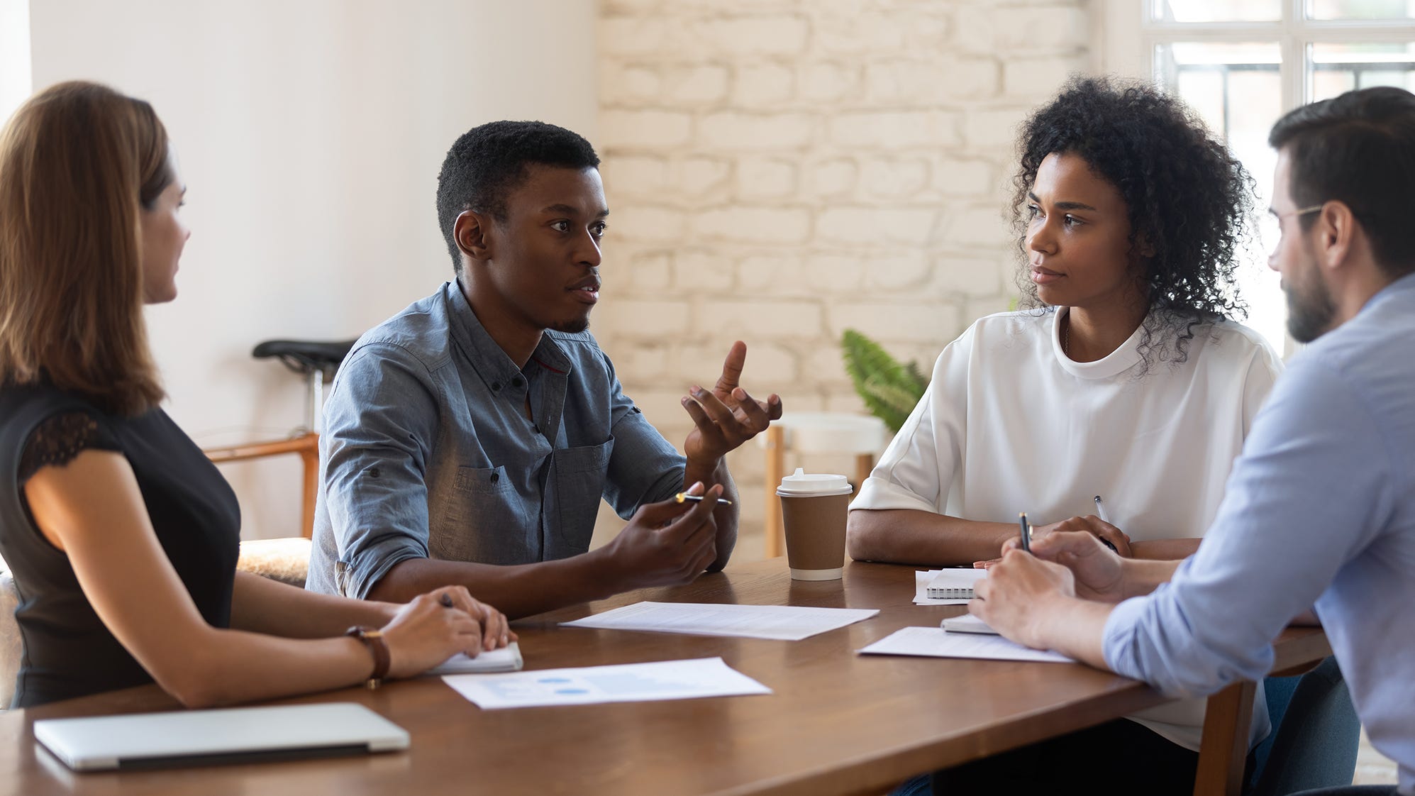 Quatro pessoas em uma mesa durante uma reunião. Uma mulher e um homem brancos, um homem negro falando e uma mulher negra.