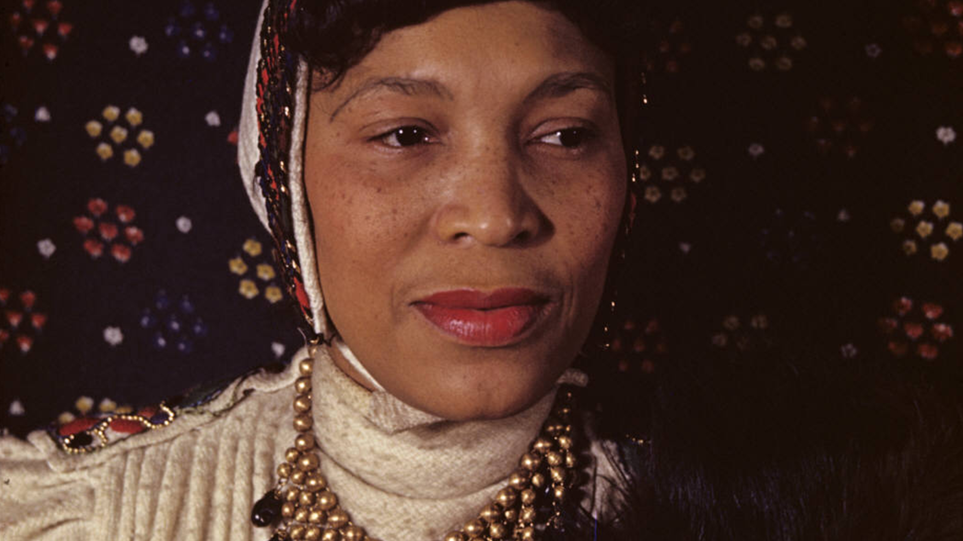 Head and shoulders portrait of Zora Neal Hurston in front of a geometric print fabric background and wearing a decorative beaded bonet
