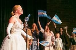 A scene from Evita in which Eva Peron stands before a microphone while her supporters wave flags in the background.