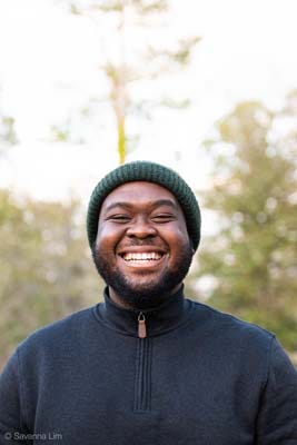 Uyiosa Elegon wears a knit cap and a fleece jacket and stands smiling at the camera for a portrait.