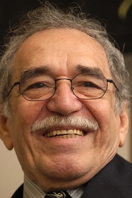Neck up picture of author Gabriel Garcia Marquez, with gray hair, a well trimmed moustache, and glasses. He’s smiling warmly.