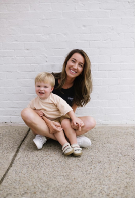 Allie Schmidt sitting on the sidewalk with her legs crossed, her son sitting on her lap. White brick wall background.