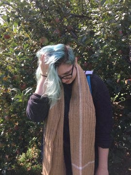 A photo of the author. She’s looking down at the ground and has one hand scrunching up in her hair (dyed blue). She’s wearing glasses, a long brown and white scarf, and a dark purple sweater. She has a blue and white bag slung over one shoulder. She’s standing in an apple orchard.