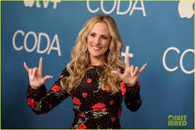 Marlee Matlin stands in front of a CODA step and repeat banner in a floral dress signaling “I love you” with both hands.