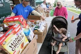 man pointing at baby in stroller and baby pointing back, man behind table full of donated food