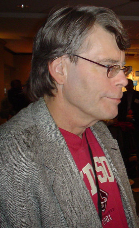 Author Stephen King, in this photo a middle aged man with slightly graying brown hair, glasses, a gray blazer over a red sports t-shirt, and a lanyard around his neck. He is in profile and appears to be mingling in a crowd of people at an event.