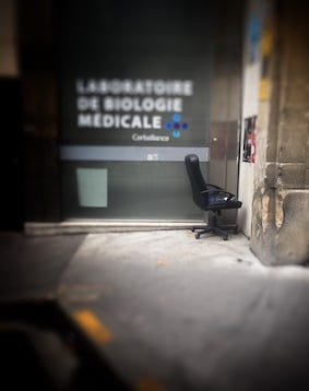 Fauteuil de bureau tourné vers la droite face à un mur. À l’arrière plan, la vitrine d’un laboratoire de biologie médicale.