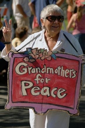 A woman with grey hair flashing a peace sign and holding a decorated sign that says: “Grandmothers for Peace”.