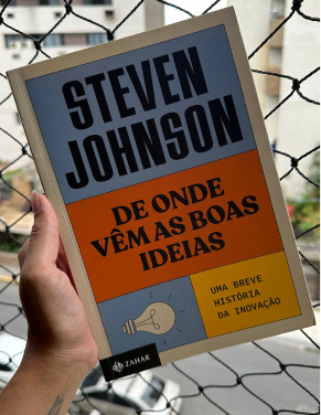 Na imagem, seguro com a mão esquerda o livro “De onde vêm as boas ideias: uma breve história da inovação”, do autor Steven Johnson. Estou segurando na janela e de fundo, tem-se uma tela de proteção de janelas e prédios da rua.