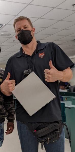 High school teacher wearing a black polo with a tiger logo on it. He has a white binder hanging from his neck and a black fanny pack around his waist. He is giving two thumbs up.