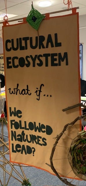 Showing an orange canvas banner with the writing ‘cultural ecosystem — what if we followed nature’s lead’ stitched on in black fabric. Banner created by Time Rebels Ruth and Odette of We Are Makers