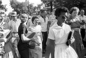 Elizabeth Eckford, a Black woman, and one of the Little Rock Nine, walking ahead of a white crowd, with a white woman screaming behind her.