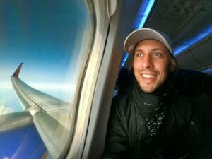 smiling man in black jacket wearing blue cap looking out airplane window.