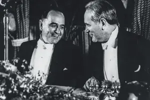 Nesta fotografia em preto e branco, dois homens (Oswaldo Aranha e Vargas) mantêm uma interação agradável em uma mesa de jantar, ambos elegantemente vestidos com smokings. Vargas, à esquerda, está sorrindo e direcionando seu olhar para a direita, em direção ao Aranha, criando uma sensação de camaradagem. Eles estão sentados em um ambiente que inclui vários talheres completos com taças de vinho e possíveis louças ou decorações, sugerindo uma ocasião de jantar formal.