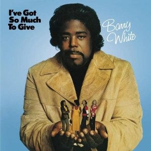 The single cover for ‘I’ve Got So Much to Give’ by Barry White. The cover is blue, with Mr. White stood in the middle wearing a cream-coloured suede leather jacket. He has black hair and a beard and moustache. In his hands are the images of four women, which he is holding out whilst looking to camera.