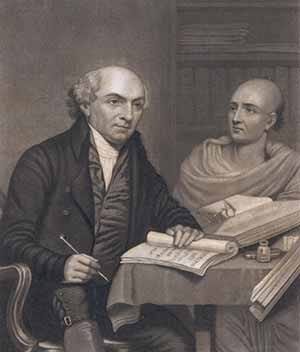 historical black-and-white engraving of a European scholar and an Indian man in traditional attire seated at a table. The scholar, holding a pen, is writing on a scroll, while the Indian man observes. Open books and ink bottles are scattered on the table, suggesting a scene of translation or study.