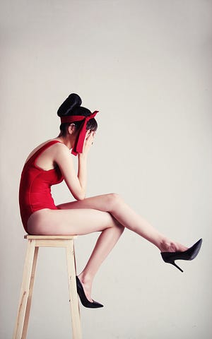 A woman in a red leotard with hands over face in despair sitting on a wooden stool.