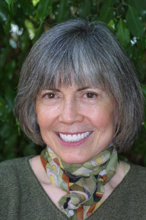 Professional looking book jacket photo of Anne Rice, in this picture an older woman with gray hair, a patterned ascot-like adornment around her neck, and green leaves in the background. She’s smiling widely and has strikingly white teeth.