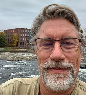Selfie image of man with glasses, grey hair, mustache and beard, River and factory in background