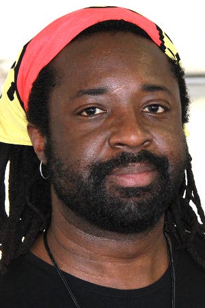 Neck up picture of author Marlon James, sporting a stubble beard, gold hoop earring, a pink headband, and long dreads. He’s also wearing a black shirt and possibly a lanyard around his neck. He has a neutral expression.