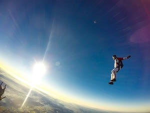 A wide-angle shot of a skydiver free falling before pulling out his parachute, set against a clear blue sky and a glaring sun