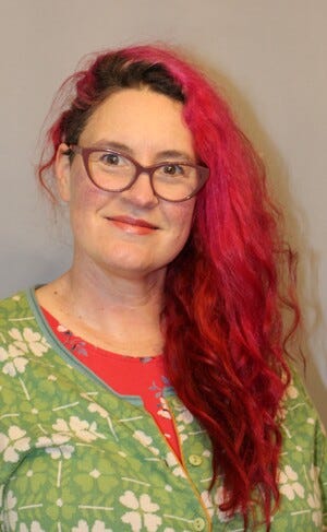 Author Image: The image of the author who is of white European ancestry, middle aged, with bright red hair, dark eyes, a soft smile and she is wearing a green floral shirt, framed glasses that have a slightly pointed ‘cat eye’ shape.
